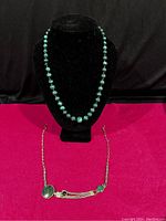 Photo shows a black velvet bust stand holding the graduated malachite bead necklace. The necklace has green malachite beads graduated from smaller to larger at the center. A small chip is visible on one bead.