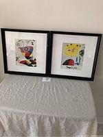 Two framed Joan Miro prints placed side by side on a covered table showing wear to black frames