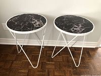 Two round side tables with black marble-patterned tops and white metal hairpin legs against a plain wall on parquet floor.