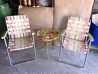Front view of two vintage folding chairs with woven pink and cream webbing and a small round floral folding table between them.