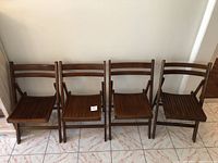 Four vintage wood folding chairs in a row against a wall on tiled floor. Three chairs match, one chair has a different slightly curved backrest design. Chairs show wear to finish.