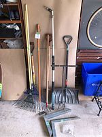 Garage floor with multiple yard tools including rakes, shovels, a spray wand, and steel L brackets, arranged upright against the wall beside a bike and recycling bin