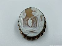 Front view of antique cameo brooch showing woman walking down a street with houses and trees.