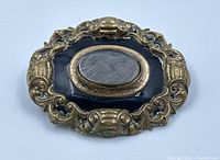 Front view of antique Victorian mourning brooch showing gold-toned ornate frame, black enamel background, and central oval with braided hair under beveled glass.