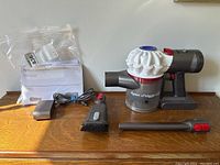 Full set of vacuum and included attachments on wooden surface showing the vacuum, charging adapter, round brush attachment, and crevice tool.