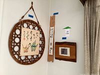 Photo showing three Japanese hanging art pieces on a wall: 18x18 paper cranes framed in circular bamboo, a 23x4 narrow strawberry art strip, and a 9x10 wooden frame with leaf fabric art.