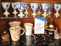 Wide view showing two shelves with Japanese ceramic and glass tea cups, mugs, and containers including blue and white goblets and decorative ceramic mugs.