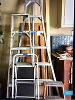 Photo showing four ladders stacked together: one large aluminum ladder, two medium step ladders, and one small foldable step ladder with black rubber steps.