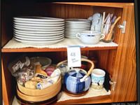 Top shelf: stacks of dinner and salad plates with handled cups; bottom shelf: two teapots and souvenir mug