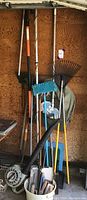 Photo showing various lawn tools such as rakes, shovels, hoes leaning against a wall with a power blower and paint tools bucket in front.