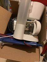 White drip coffee maker with a glass carafe inside a cardboard box, showing it is packed with other items