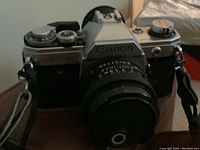 Front view of Canon AE-1 camera with fixed lens and attached strap, showing Canon branding and lens cap.