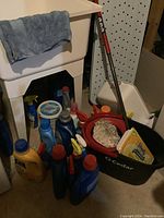 Photo showing a basement corner with a white plastic utility basin sink and grey towel, multiple bottles of laundry detergent and cleaning sprays grouped nearby, black O-Cedar mop bucket with a string mop inside.