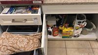 View of open kitchen drawers and cabinet containing liquid soaps, plastic wraps, paper napkins and kitchen storage items
