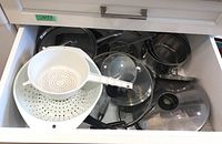 Drawer containing stainless steel sauce pans with black handles, various glass lids with black knobs, and two white plastic strainers/colanders