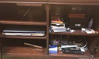 Wooden shelving unit with various electronic items including a laptop, remote controls, cables, and small accessories.