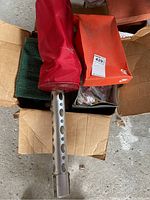 Box containing a red golf club cover with silver metal ventilation holes, an orange golf club cover, and possibly a towel in a rough cardboard box in a garage.