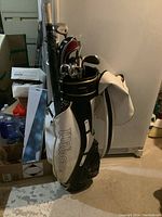 Photo showing Wilson branded golf bag filled with golf clubs and cover, set stored in basement area with boxes and other items around