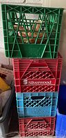 Stack of four assorted plastic milk crates in green, red, and light blue, showing front branding and general condition needing cleaning.