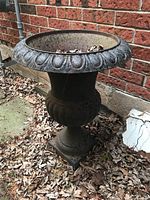 Front and side view of the tall cast iron planter showing its urn shape and decorative lip, with visible rust and soil inside with dried leaves.