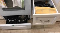 Open drawer showing pots and pans inside, some with lids, and folded yellow and cream colored cloths.