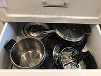 Drawer containing assorted cookware including a large stainless steel pot with lid, non-stick skillets, a whisk, and disposable pie plates.