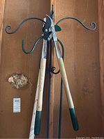 Photo showing branch lopper with wooden handles and metal cutting blades hanging on one shepherds hook, part of the shepherds hooks visible with curved ends