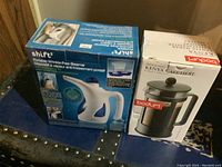Shift portable steamer and Bodum Kenya French press boxed products positioned side by side on a surface.