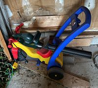 Plastic child's trike in yellow, red, blue, and green plastic seen in garage against a wall with dirt on floor.
