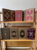Nine leather-bound books arranged on two shelves, showing full gilt-decorated covers