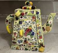 Front view of the ceramic teapot-shaped wall cabinet showing colorful mosaic fruit and bee patterns, and 3D fruit accents on handle and top.