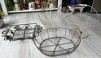 Four wire baskets displayed on floor showing variety of styles and shapes including large round basket, smaller baskets, and decorative trivet stand.
