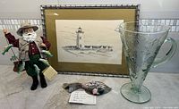 Kurt Adler Santa fisherman figure holding fish on multiple fishing lines, standing on table next to a green glass pitcher embossed with sea shells and a framed black-white lighthouse print. Porcelain fish artisan piece placed in front of the framed print.
