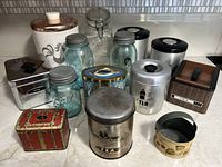 Overview of all cans, canisters, jars, and tins displayed on a countertop. Includes a mix of metal, glass, ceramic pieces with various labels and designs showing vintage usage.