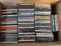 Photo showing approximately 90 assorted music CDs stacked in a box featuring rock, jazz, and indie genres with visible album spines.