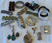 Photo showing full collection of costume jewelry bracelets and pendants spread on a fabric surface. Multiple bracelets in silver-tone, rhinestone, green stones, and black and white bangles. Several pendants including Brighton and 925 stamped pieces.
