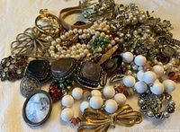 Photo showing a pile of mixed broken costume jewelry including faux pearl strands, white beads, various brooches, and pendants with worn metal and stones.