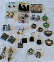 Photo showing 18 pairs of pierced costume jewelry earrings arranged on a white textured background, including branded items from 1928, Jennifer Moore, Sarah Coventry, Smithsonian Institute, and Chico's.