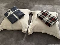 Two large Euro pillows with cream-color Martha Stewart shams visible, laid on carpet. Navy blue throw blanket in packaging on left pillow, plaid throw folded on right pillow