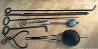 XL oversized black metal fireplace tools including poker, tongs, curved poker, popcorn popper, and ice tongs arranged on floor.