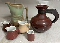 Photo showing Frankomia pitcher in green with bamboo-style handle, Franciscan Ware California brown and black carafe with wooden handle, and four Hall creamers in small to medium sizes with various brown and beige glazes.
