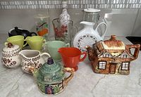 Various vintage and contemporary pitchers, creamers, decorative teapots, and jars arranged on a marble countertop showing diverse styles and materials including ceramic and glass.