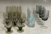 Image of 5 small smokey grey conical aperitif/dessert glasses, 8 taller smokey grey glasses, 4 grey wine glasses, and 2 decorated turquoise/gold glasses arranged on a surface.