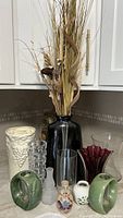 Full view of all vases arranged on kitchen counter including large black ceramic vase with dried flowers, cream ceramic vase, green ceramic vases, maroon glass vase, crystal glass vases, and small decorative vases.