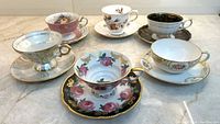 Shows six vintage tea cups and saucers arranged on a surface featuring different floral and metallic designs with varying colors like pink, black, grey, and white.