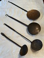 Four vintage metal kitchen utensils arranged on white cloth, including various perforated spoons and ladle with long handles