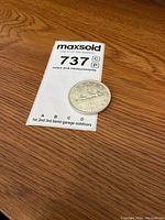 Silver Canadian dollar coin placed next to an auction tag showing lot number 737 on a wooden surface.
