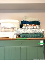Nine folded blankets stacked in two piles on a green cabinet, showing various textures and colors including white, cream, teal, and patterned blankets.