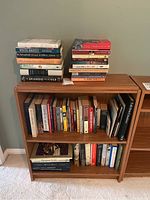 Photo showing two stacked piles of books on top of a wooden bookshelf filled with multiple books, various titles and authors visible