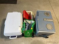 Three coolers including a large white/blue cooler, a smaller cream/red cooler, and a blue Mobicool electric cooler alongside a green reusable bag containing ice packs.